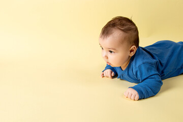 A beautiful little baby in a blue bodysuit, lies on the stomach on a yellow background. Concept, mockup, blank, copy space.