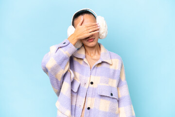Young Arab woman wearing winter muffs isolated on blue background covering eyes by hands. Do not...