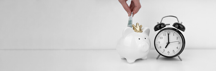 Womans hand put dollar bancnote into a piggy bank with alarm clock