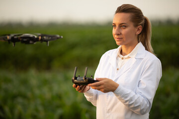 female agricultural specialist holding Drone Remote and controlling drone in air standing in corn...