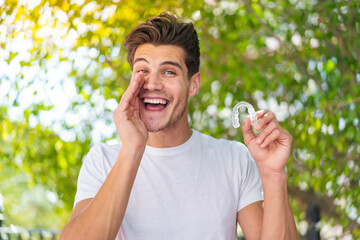 Young caucasian man holding invisible braces at outdoors shouting with mouth wide open