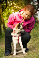 Woman hugging dog in forest. Cheerful lady in pink jacket hugs puppy sitting on a path of the forest on nice day.