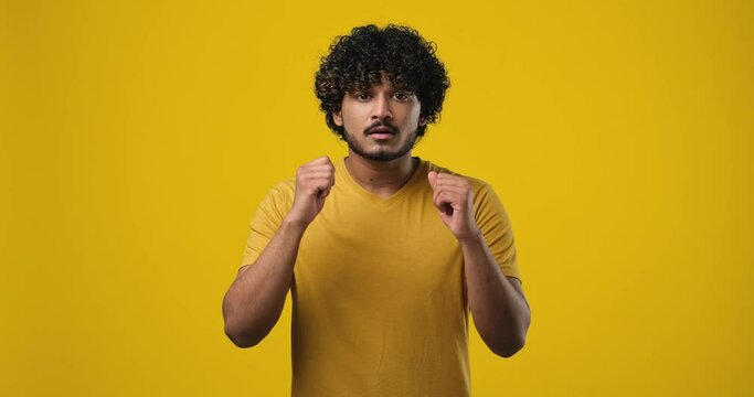 Young indian man watching game with fingers crossed for luch and celebrating victory enjoying success standing over orange studio backround