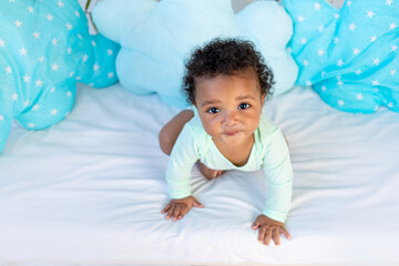 cute african baby sitting in bed at home before going to bed on a blue bed and looking up, the concept of healthy sleep and baby bedding cotton accessories - obrazy, fototapety, plakaty