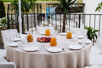 The round table is covered with a white tablecloth and served with dishes on the open terrace of the restaurant. Gourmet outdoor breakfast with fruit and orange juice.