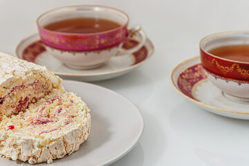 delicious meringue roll with strawberry cream and vintage cups of tea on a white glass surface