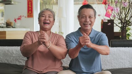 Medium shot of Asian senior family couple sitting on couch in living room decorated for lunar New Year, looking at camera, holding hands clasped and saying wishes with smile