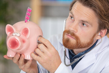 doctor holding stethoscope to piggybank continaing blister of tablets - obrazy, fototapety, plakaty