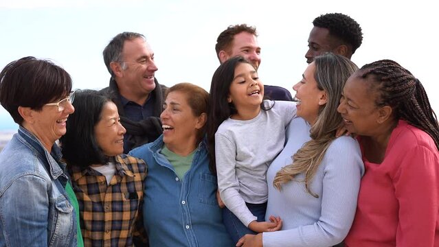 Group of multigenerational people hugging each other while smiling in front of camera - Multiracial friends having fun together outdoor
