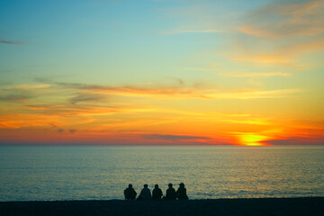 Young teenage people - guys and girls - sit on the beach and watch the sunset. Group of five happy people sits on background of empty sunset beach. Travel or sea vacations concept. back, rear view.