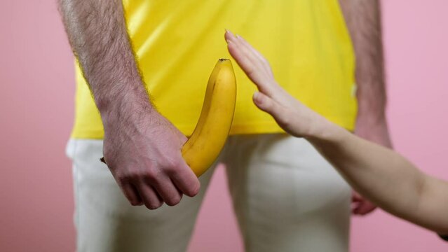 Man holds a rised banana in his hand at the level of the genitals. A woman's hand runs a finger over a banana. Pink background. The concept of men's health