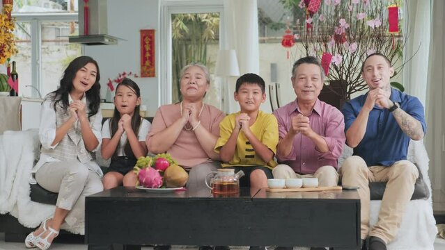 Happy large Asian family sitting on sofa in living room, looking at camera and saying wishes while celebrating Lunar New Year at home