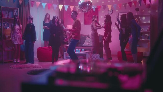 A group of friends at a party. Children dance in a room with a spinning disco ball. The girl in black feels awkward, modestly dancing next to the guy. On the table are paper cups, pizza and popcorn.