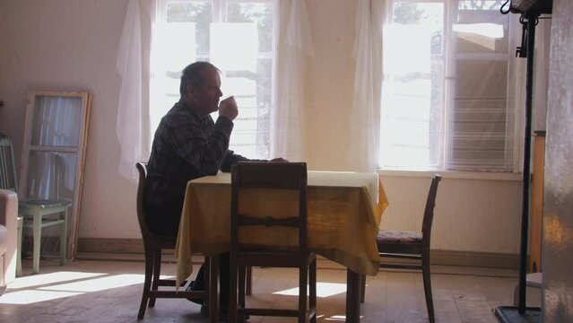 Retired elderly man, sitting alone sipping coffee at rustic table. Side profile