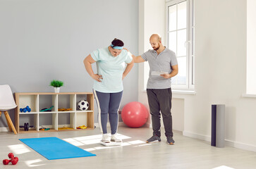 Personal trainer evaluating health of overweight young woman in gym. Instructor checking progress of plump woman, who standing on scale in fitness center. Sport, slimming, dieting