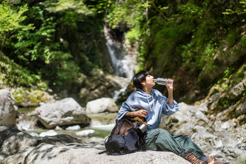 トレッキングをしている若い日本人女性/渓谷の岩に座り水を飲んでいる - obrazy, fototapety, plakaty