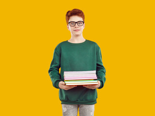 Funny redhaired schoolboy with glasses with droll surprised expression face looks at the camera, dressed in green sweater and gray jeans, holding several school notebooks on isolated yellow background