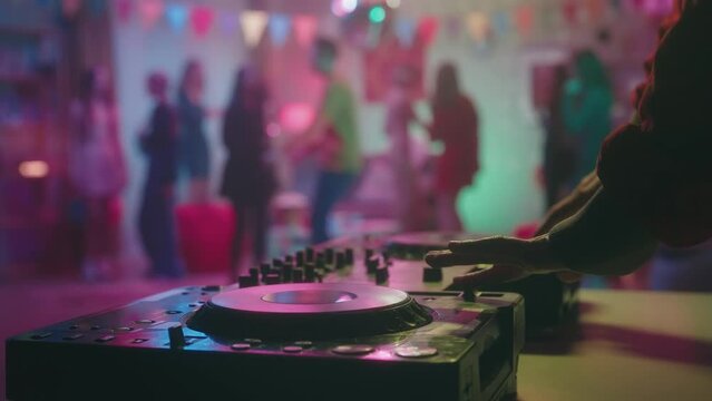 Male hands mix music on a dj console against a blurred background of dancing people. Camera focus on the dancing teenagers in a decorated party room with a disco ball. The girl in black feels awkward.