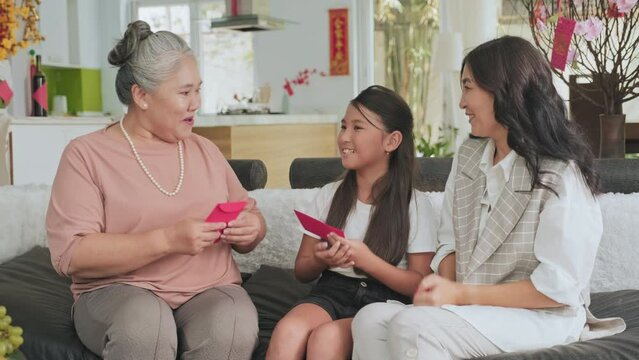 Senior Asian grandmother giving lucky money in red envelopes to joyous little granddaughter and daughter on lunar New Year celebration at home
