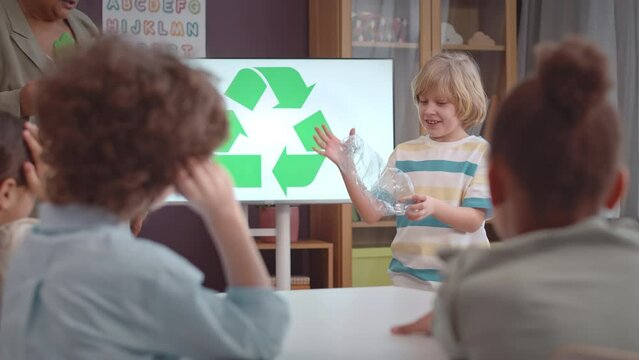 Medium shot of Caucasian first grade school boy with smashed plastic bottle in hands making presentation on recycling to teacher and classmates during Ecology and Environment lesson