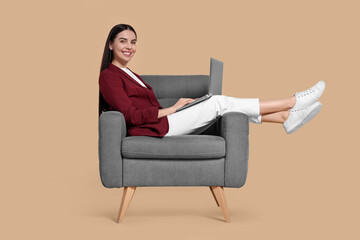 Fototapeta na wymiar Happy woman with laptop sitting in armchair on beige background