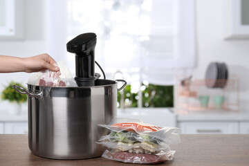Woman putting vacuum packed meat into pot with sous vide cooker on wooden table in kitchen, closeup. Thermal immersion circulator - obrazy, fototapety, plakaty