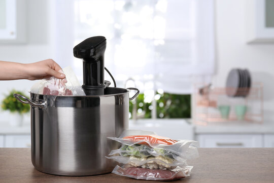 Woman putting vacuum packed meat into pot with sous vide cooker on wooden table in kitchen, closeup. Thermal immersion circulator