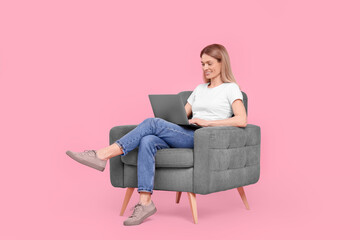 Fototapeta na wymiar Happy woman with laptop sitting in armchair against pink background