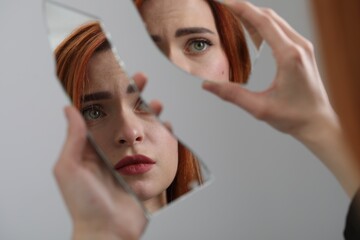 Young woman looking at herself in shard of broken mirror on light grey background, closeup - obrazy, fototapety, plakaty