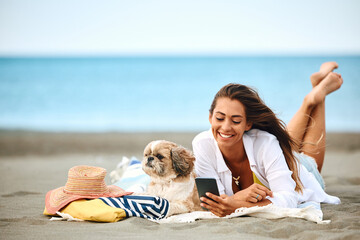 Happy woman texting on cell phone while relaxing with her dog on beach.