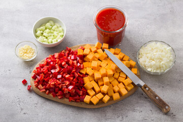 Wooden board with chopped sweet peppers and pumpkin, chopped onion, celery, garlic and tomato sauce on a gray textured background, top view. Cooking vegan food