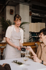 Cheerful young blonde waitress accepts payment through bank terminal for lunch of happy mature woman in cafe