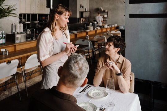 Concentrated young blonde waitress writes down order of positive couple in cafe