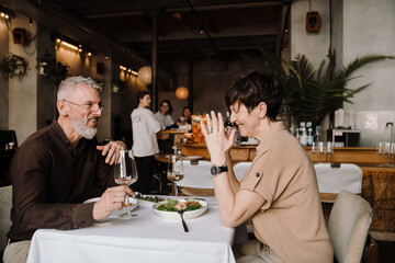 Cheerful mature couple dining with glasses of wine and talking in cafe