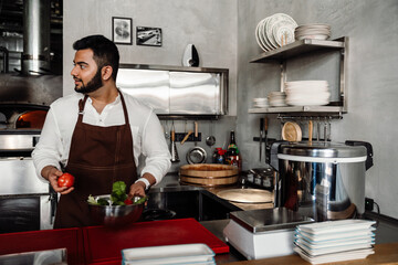 Pretty young indian chef in apron looking to side and going to make salad in cafe kitchen