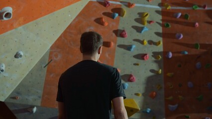 A millennial male climber with his back to the camera walks towards a climbing wall with...