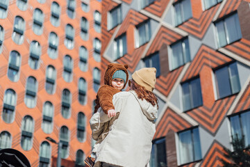 a woman from the back holds a child in a teddy bear hoodie in her arms against the background of a modern building in the city