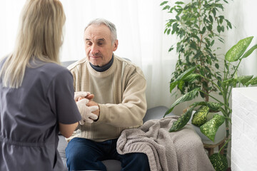 Caring nurse talks to old patient holds his hand sit in living room at homecare visit provide...