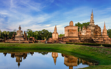 Fototapeta na wymiar Beautiful wide view of ruins of Wat Mahathat Sukhothai at sun set with white clouds and blue sky on background. UNESCO and World Heritage Site. Travel Concept.
