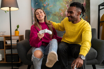 Cheerful multiracial boyfriend and girlfriend in colorful casual clothes sitting on sofa with gamepads and playing video game together in modern living room