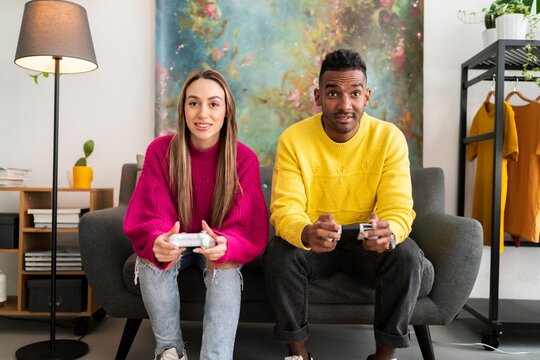 Cheerful multiracial boyfriend and girlfriend in colorful casual clothes sitting on sofa with gamepads and playing video game together in modern living room