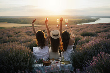 Silhouettes of three happy young girls with hands up sitting and drinking wine on beautiful sunset.