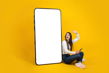 Woman sitting on the floor pointing finger on huge blank screen mobile phone mockup area. Holding using smartphone. Full body length happy smiling cheerful girl recommending application. Copy space.