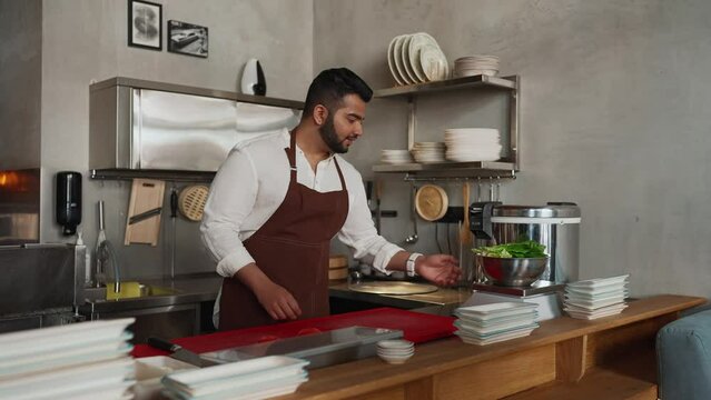 Concentrated indian chef in apron weighs lettuce leaves in cafe kitchen