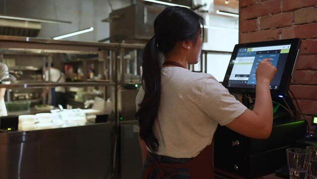Back view of brunette asian business owner in apron punches food order on cash register in cafe