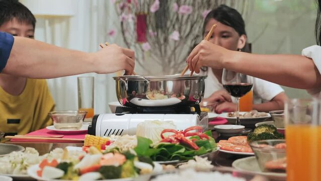 Mother and dad using chopsticks for putting food ingredients in hot pot while cooking traditional Vietnamese dishes on Tet holiday dinner with kids