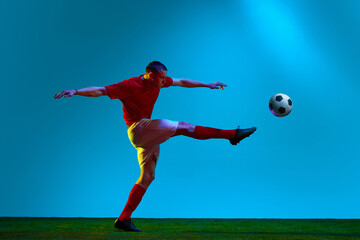 Fototapeta na wymiar Portrait of soccer, football player in sports uniform kicking the ball in motion over field background in neon light. Hitting ball with leg