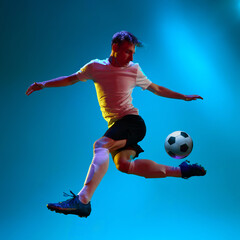 Dynamic portrait of soccer, football player in sports uniform kicking the ball over blue background...