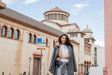 Fototapeta premium Smiling and brunette woman in casual jacket holding fresh orange and leash while standing near blurred historic landmark on urban street in Barcelona, Spain, ancient building