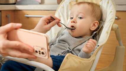 Portrait of cute baby boy eating porridge in highchair and watching cartoons on smartphone